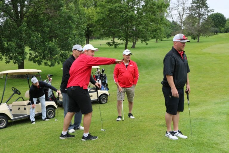 A group of golfers in casual attire, discussing a shot near golf carts on a lush green course.