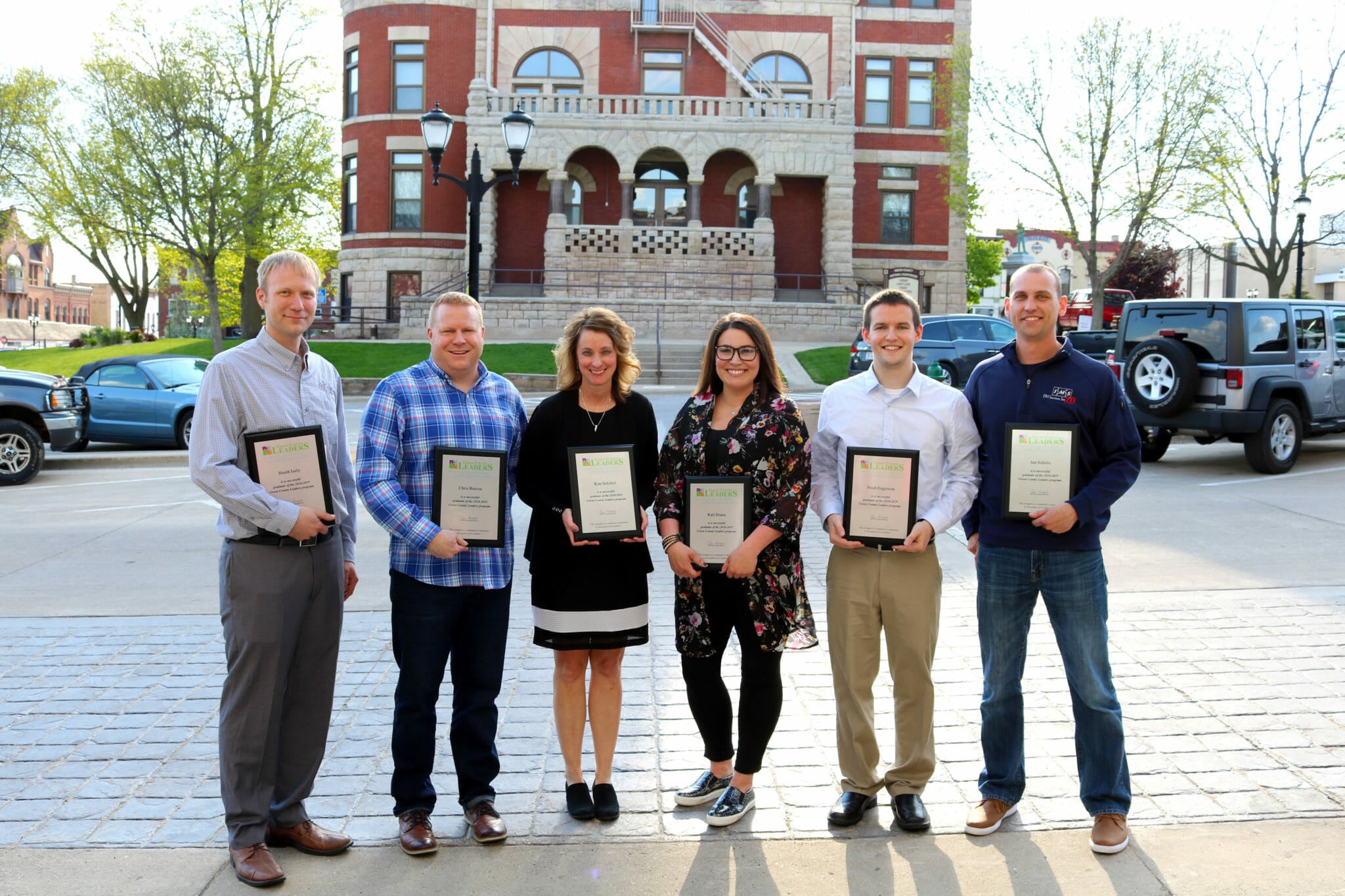 Six individuals hold awards outside a historic building, with trees and parked cars in the background.