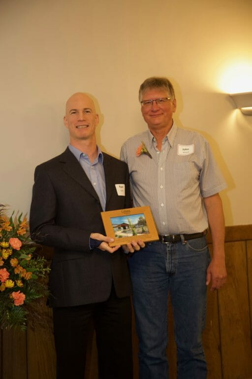 Two men pose together, one holding a framed photo, with a floral arrangement in the foreground and a warm, indoor setting.