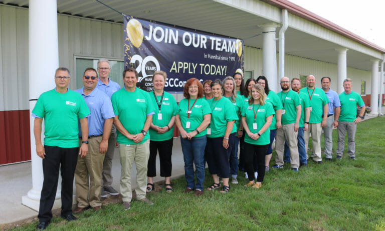 Group of employees in green shirts standing in front of a building, promoting job openings with a banner above.