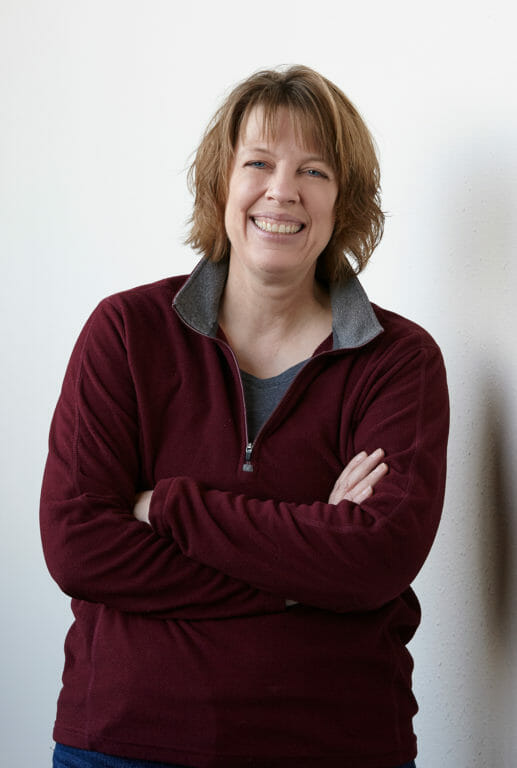 Smiling woman with shoulder-length hair, wearing a maroon zip-up fleece, stands with arms crossed against a white wall.