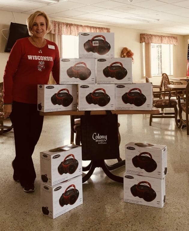 A woman in a Wisconsin shirt stands beside a stack of boxed portable CD players in a bright, communal space.