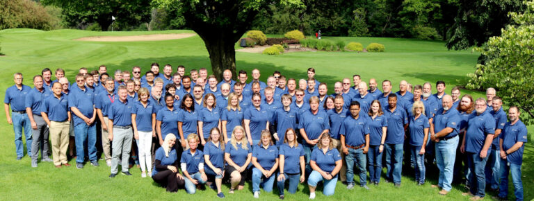 A large group of diverse individuals in matching blue shirts poses together on a lush green lawn, with trees in the background.