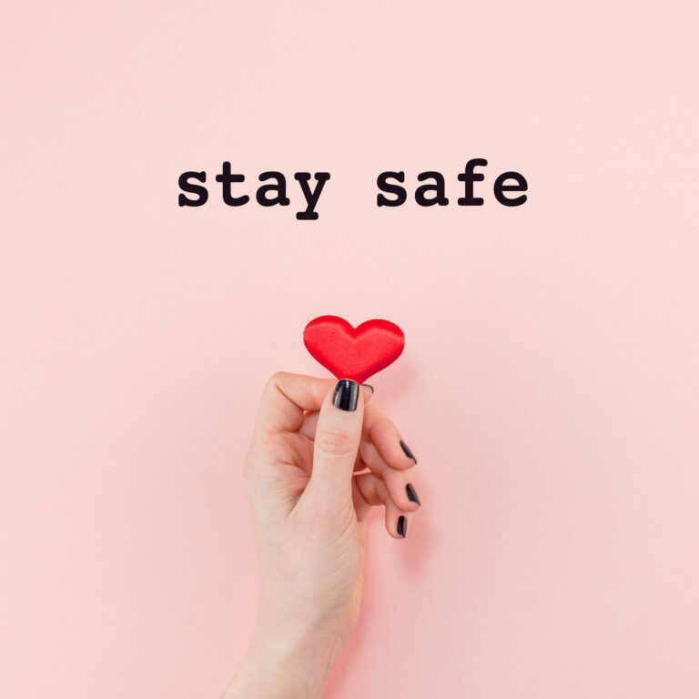 A hand holding a small red heart against a soft pink background with the text "stay safe" above.