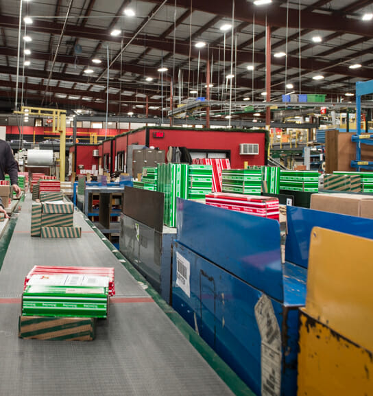 Conveyor belts lined with colorful boxes in a busy warehouse, with workers and machinery in the background.