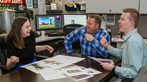 Three colleagues laugh and discuss design plans at a conference table, surrounded by sketches and computer screens.