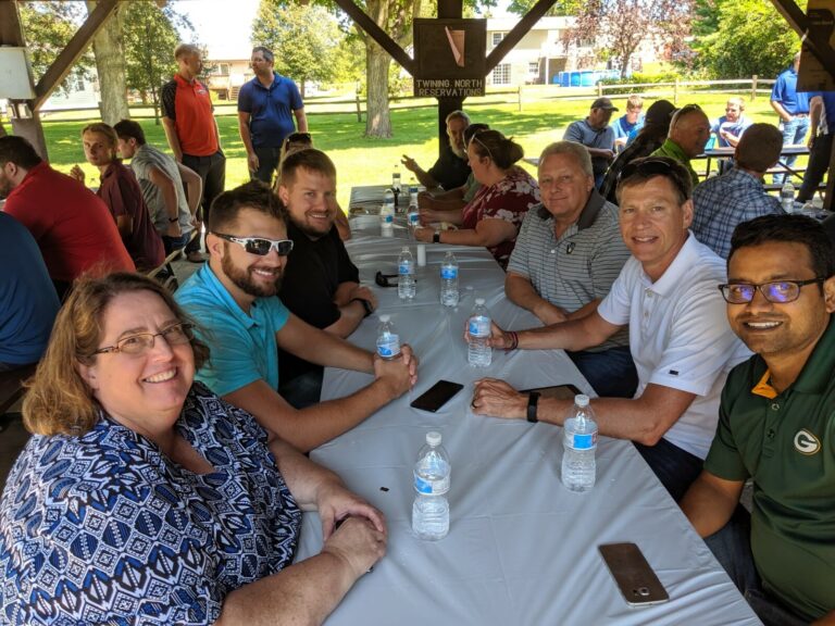 Group of six smiling people sitting at a table with water bottles, under a pavilion in a sunny outdoor setting.