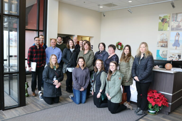 A diverse group of 15 people poses together in a modern office lobby decorated for the holidays.