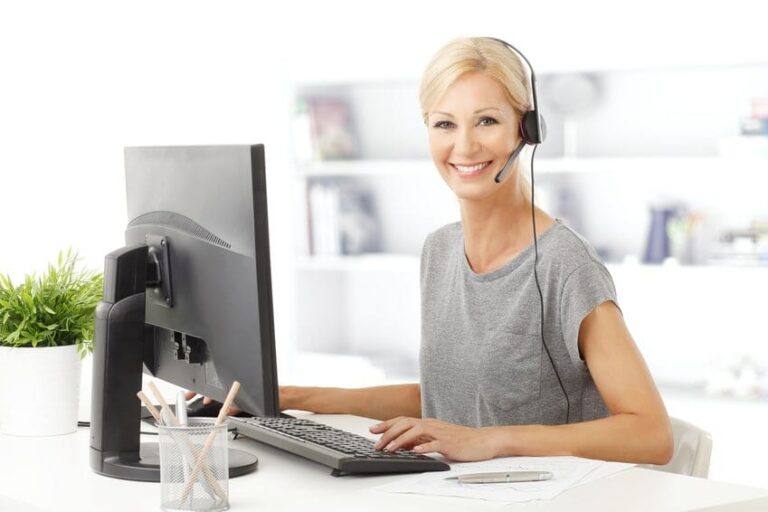 Smiling woman with a headset sits at a desk, working on a computer, surrounded by a plant and office supplies.