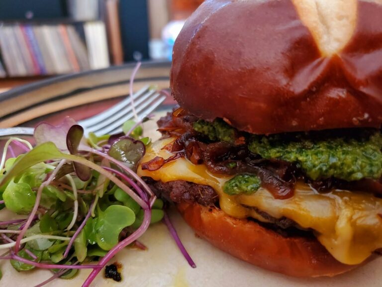 A close-up of a pretzel bun burger with melted cheese, caramelized onions, and green pesto, accompanied by a fresh microgreens salad.