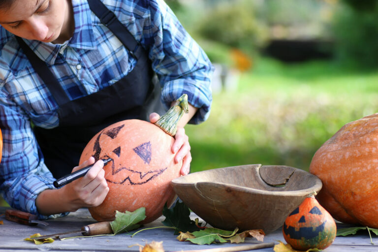 A person draws a jack-o'-lantern face on a pumpkin, surrounded by leaves and a wooden bowl, in a sunny outdoor setting.